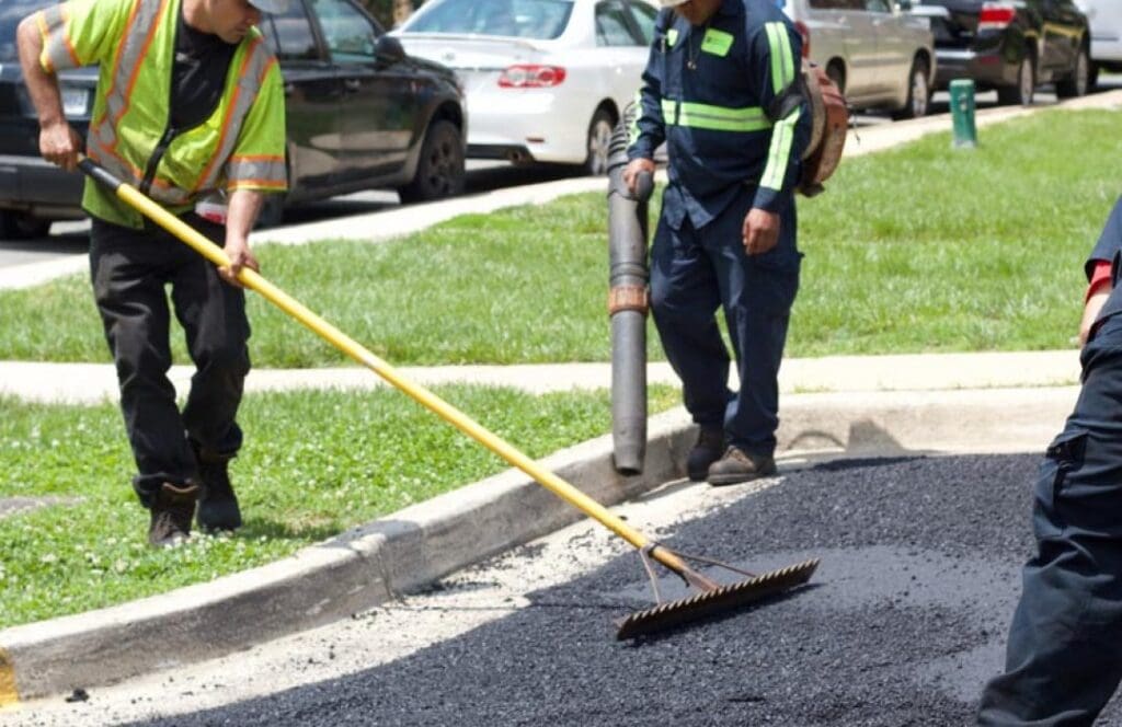 Recycled asphalt being applied along a curb by construction workers during pavement maintenance.